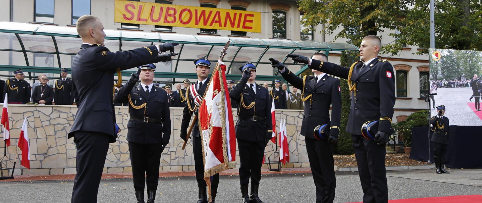 Czterech kadetów składa uroczyste ślubowanie na sztandar Szkoły, który trzyma poczet sztandarowy, w głębi na trybunie goście uroczystości, w tle budynek z przywieszonym banerem z napisem ŚLUBOWANIE