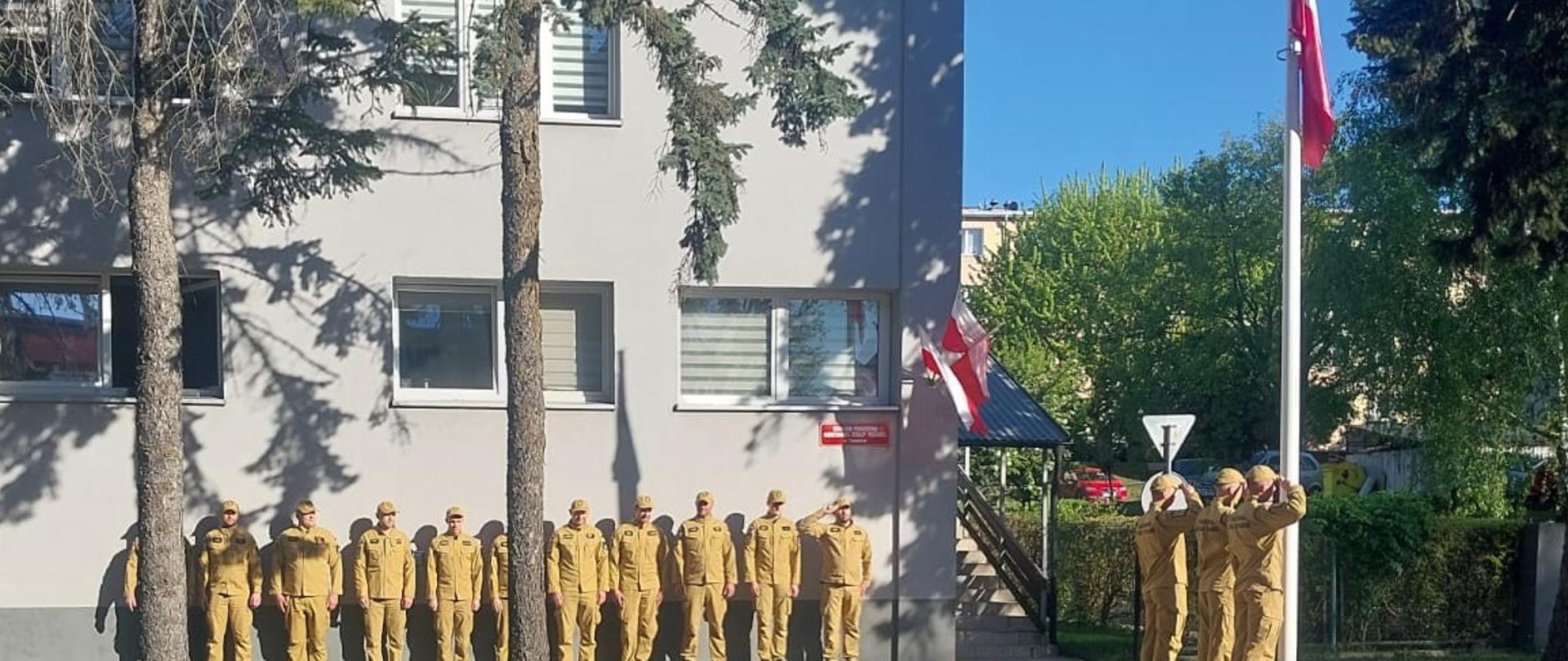 Grupa osób w jednakowych, żółtych mundurach stoi w szeregu przy ścianie jasnego budynku. Po prawej stronie trzy osoby salutują przy maszcie, na którym wisi biało-czerwona flaga. Wokół znajdują się drzewa, trawnik i chodnik, a scena wygląda jak oficjalna zbiórka lub ceremonia na świeżym powietrzu.