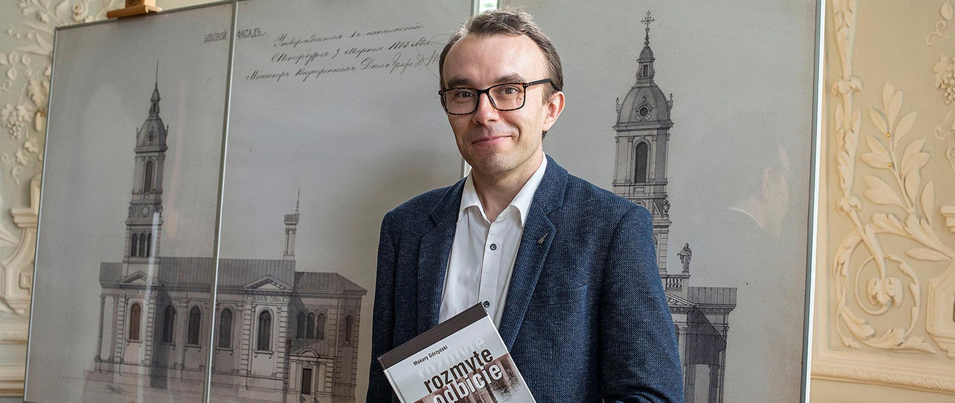 To zbliżenie na młodszego mężczyznę w okularach i niebieskiej marynarce, który trzyma książkę „Rozmyte odbicie”. Stoi on na tle trzech dużych tablic wystawowych ustawionych na sztalugach. Na tablicach widnieją precyzyjne szkice architektoniczne elewacji i przekrojów klasycystycznego budynku z wysoką wieżą. Wnętrze jest jasne, oświetlone naturalnym światłem wpadającym przez okno, a ściany zdobią delikatne, wypukłe ornamenty roślinne.