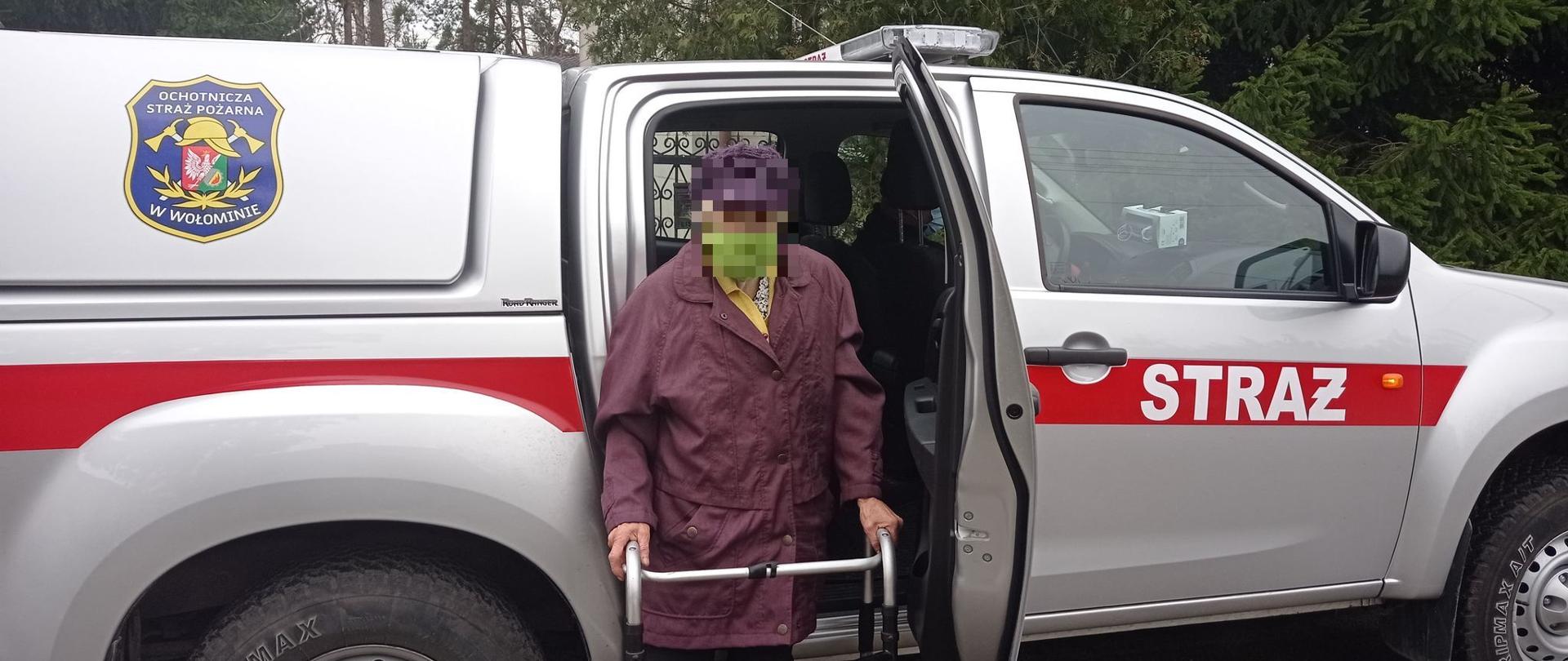 Na pierwszym planie na zdjęciu widać samochód OSP typu pic-up. Do środka wsiada kobieta, która ma założoną maseczkę na twarzy. 