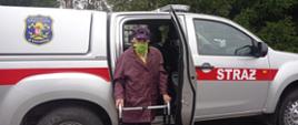 Na pierwszym planie na zdjęciu widać samochód OSP typu pic-up. Do środka wsiada kobieta, która ma założoną maseczkę na twarzy. 