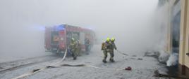na placu przed budynkiem panuje duże zadymienie, strażacy rozwijają linie gaśnicze
