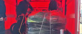 Zdjęcie przedstawia paletę z ubraniami NOMEX, która znajduje się na pace samochodu kwatermistrzowskiego. Na zdjęciu widać również strażaków