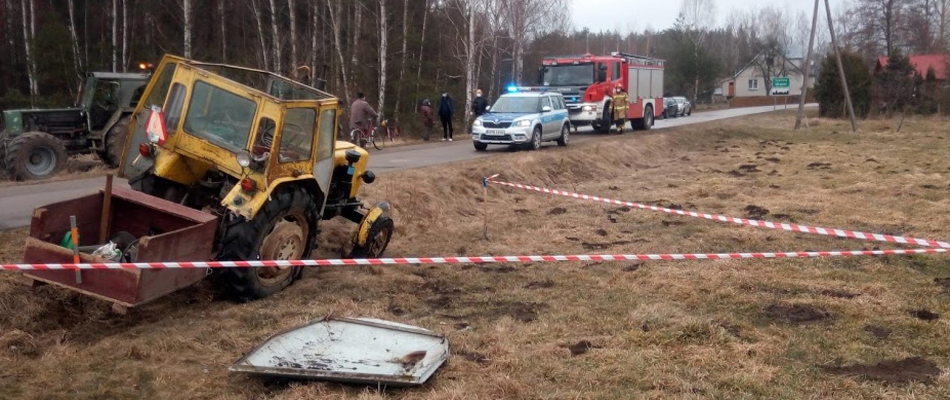 Na zdjęciu widać przechylony ciągnik rolniczy z rozbita kabiną w pobliskim rowie, ogrodzony biało czerwoną taśmą ostrzegawczą 