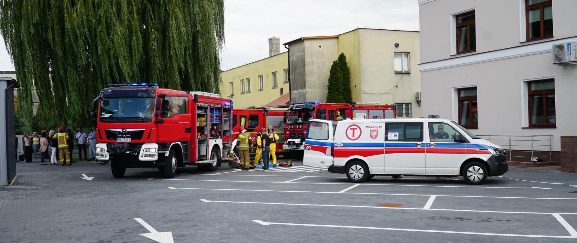 Zdjęcie przedstawia plac manewrowy lub parking przed nowoczesnym, jasnym budynkiem, na którym trwa akcja ratownicza lub ćwiczenia. Centralnie stoi duży, czerwony wóz strażacki marki MAN z włączonymi niebieskimi sygnałami świetlnymi i otwartymi skrytkami bocznymi. Obok niego, tyłem do obiektywu, stoi biały furgon ratowniczy Volkswagen Transporter z otwartą tylną klapą. Na boku furgonu widoczne jest logo (litera T w kole) oraz napis "Mazowsze". W głębi widać kolejny wóz strażacki oraz grupę kilku strażaków w żółtych mundurach bojowych. Po lewej stronie wierzba płacząca, a w oddali widać cywilów obserwujących zdarzenie. Nawierzchnia placu wykonana jest z ciemnej kostki brukowej z wymalowanymi białymi liniami parkingowymi.