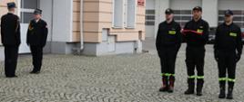 Trzech strażaków stoi trzymając flagę Polski obok funkcjonariusz Państwowej Straży Pożarnej składa meldunek zastępcy pomorskiego komendanta wojewódzkiego Państwowej Straży Pożarnej.