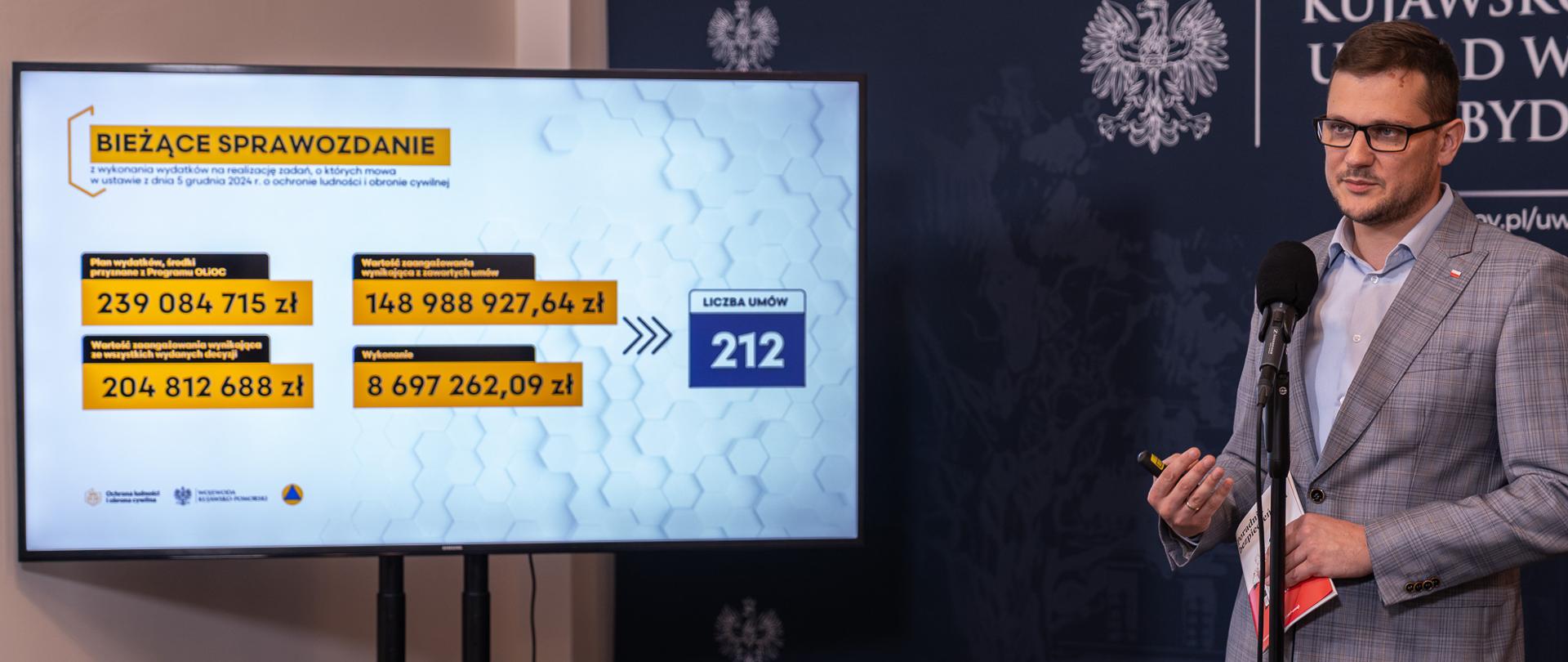 Wojewoda omawia prezentację na konferencji prasowej. Obok niego stoi telewizor, na którym wyświetlana jest prezentacja. W ręku Wojewoda trzyma Poradnik Bezpieczeństwa. Za Wojewodą stoi granatowa ścianka z logotypami urzędu wojewódzkiego.