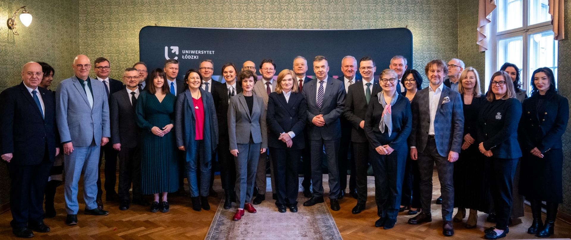 Zdjęcie zbiorowe, pod zieloną ścianą sali na podłodze z drewnianej klepki stoi duża grupa osób.