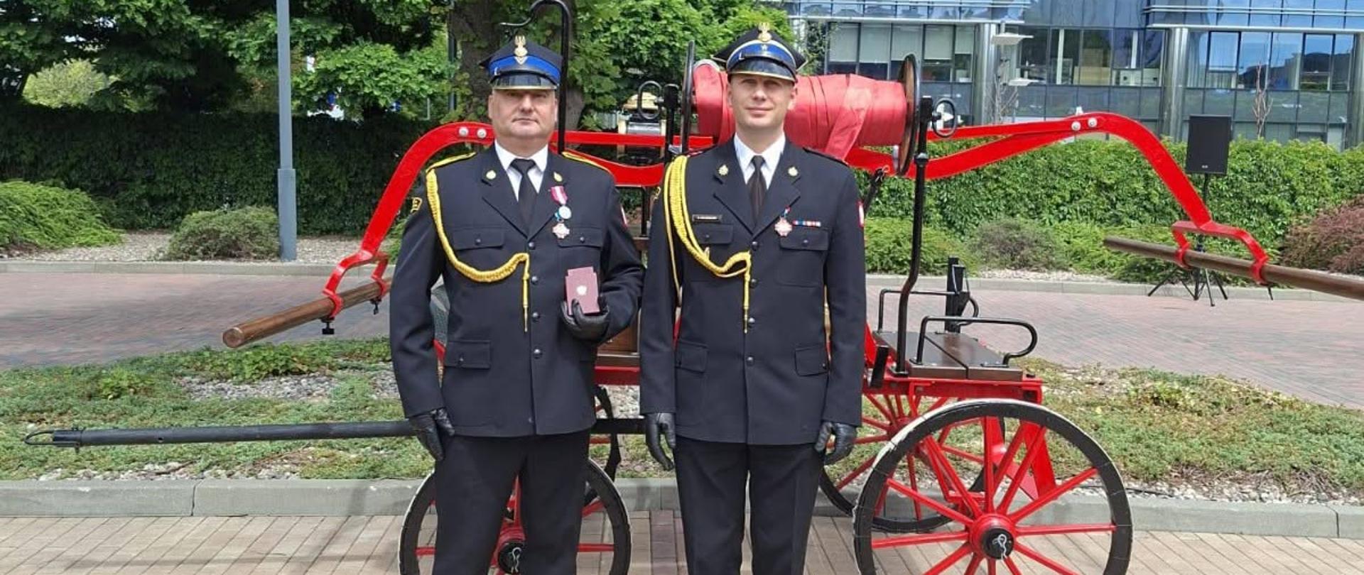 Wspólne zdjęcie wyróżnionego funkcjonariusza KP PSP w Makowie Mazowieckim z Komendantem Powiatowym na tle drzew i pojazdu strażackiego. Z tyłu widać duży przeszklony budynek. 