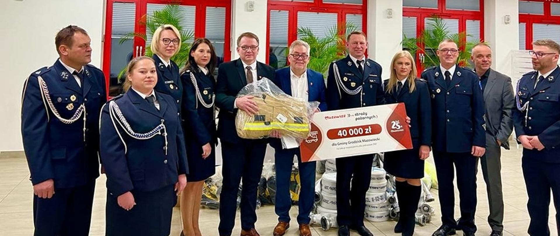 Przedstawiciele gminy Grodzisk Mazowiecki wraz z druhami i druhnami pozują na tle otrzymanych prezentów od samorządu.