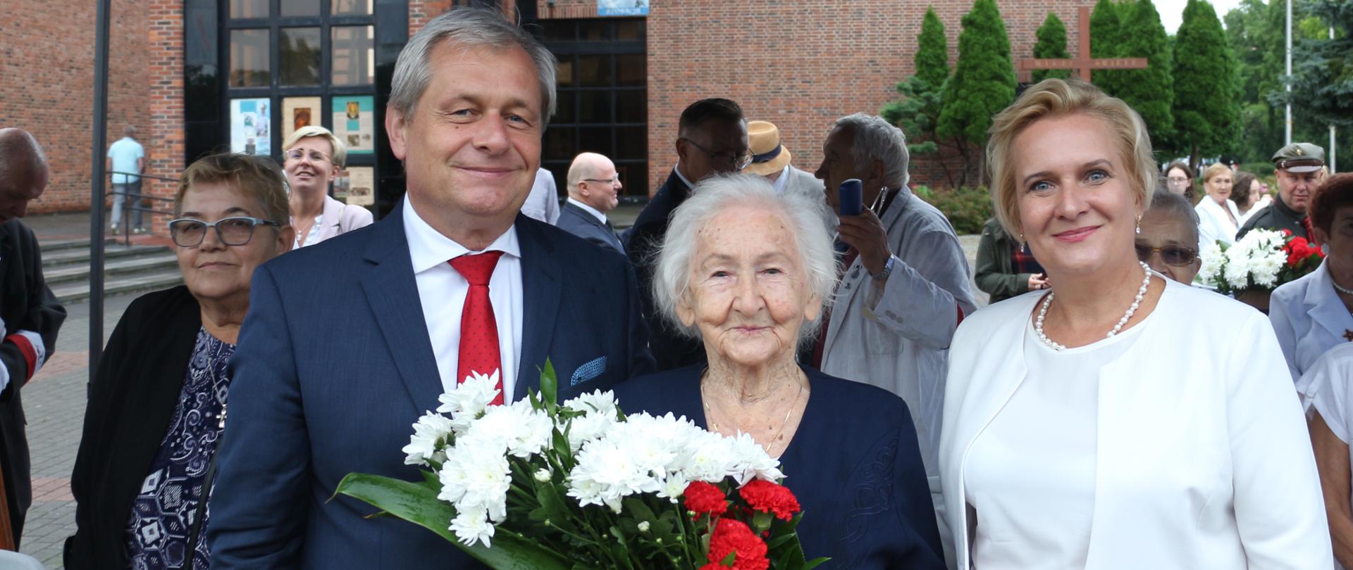 Na zdjęciu od lewej stojący w garniturze wojewoda opolski, obok niego starsza pani z siwymi włosami - to uczestniczka Powstania Warszawskiego. W dłoniach trzyma biało-czerwony bukiet kwiatów. Obok niej, po prawej stronie kadru, widać wicewojewodę opolskiego ubraną na biało-czerwono. W tle widać pozostałych uczestników uroczystości oraz fragment kościoła. Powyżej kadru zarys namiotu. 