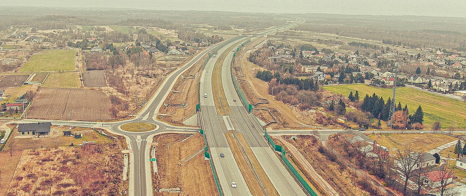 Fotografia lotnicza autostrady A1 na terenie Blachowni wykonana w listopadzie 2021 r. Na pierwszym planie widoczny wiadukt autostradowy nad rondem drogowym położnym pod jezdnią autostrady. Po obu stronach autostrady widoczna zabudowa jednorodzinna poprzecinana ternami rolniczymi i leśnymi. 