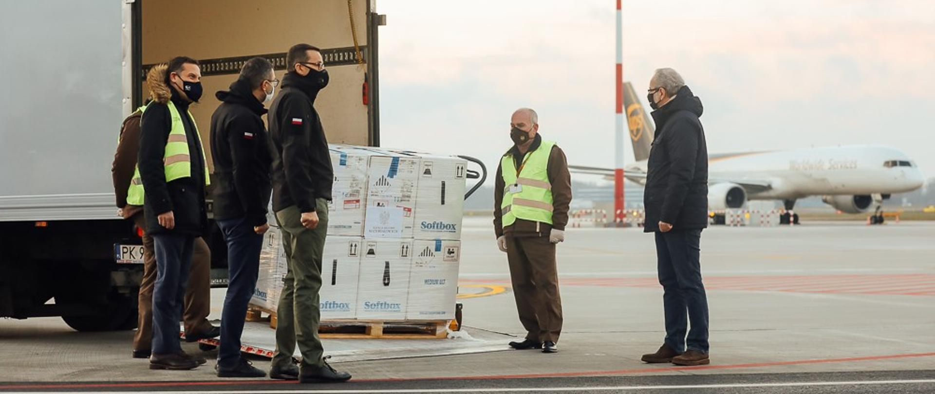Rozładunek paczek na płycie lotniska. Przesyłkę oglądają premier Mateusz Morawiecki, minister Adam Niedzielski oraz Michał Kuczmierowski.