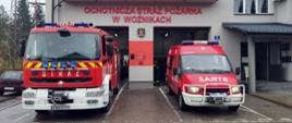 Dwa samochody pożarnicze stoją przed bramami garażowymi w remizie OSP.