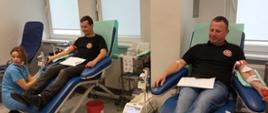 Na zdjęciu dwaj mężczyźni po lewej w czarnej koszulce i czarnych spodniach po prawej w czarnej koszulce i dżinsach siedzą na granatowych fotelach oddają krew w pomieszczeniu o białych ścianach i szarej posadzce w Regionalnym Centrum Krwiodawstwa i Krwiolecznictwa w Oddziale Terenowym w Gorlicach.