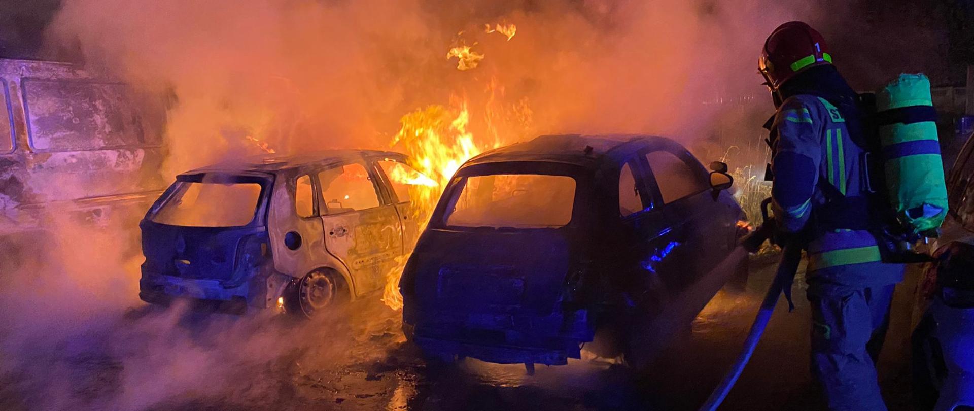 Zdjęcie wykonane jest w nocy. Na pierwszym planie widać strażaka gaszącego płonące auta. Na drugim planie widać dwa palące się auta i unoszący się biały dym. Całe zdjęcie ma niebieską poświatę.