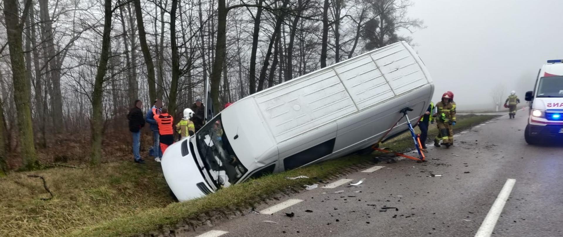 Na zdjęciu widoczny bus leżący na lewym boku w rowie, za nim ratownik medyczny oraz strażak. Po prawej stronie na drodze stoi karetka pogotowia ratunkowego.