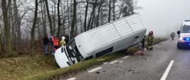 Na zdjęciu widoczny bus leżący na lewym boku w rowie, za nim ratownik medyczny oraz strażak. Po prawej stronie na drodze stoi karetka pogotowia ratunkowego.