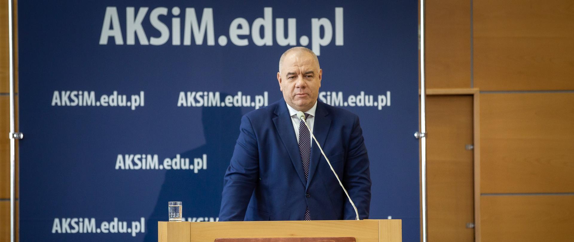 Wicepremier Jacek Sasin stoi za mównicą. W tle ścianka z adresem AKSiM.edu.pl. Ujęcie zza słuchaczy.