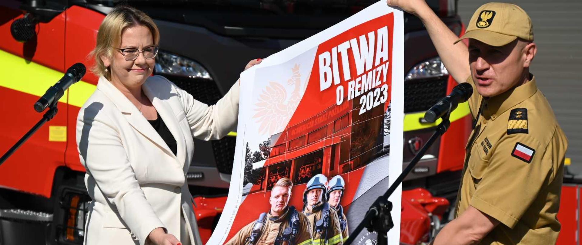 Strażak - generał w mundurze służbowym oraz pani minister prezentują we dwoje - rozwijają - plakat informacyjny. Na plakacie strażacy. W tle za nimi pojazdy pożarnicze.