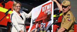 Strażak - generał w mundurze służbowym oraz pani minister prezentują we dwoje - rozwijają - plakat informacyjny. Na plakacie strażacy. W tle za nimi pojazdy pożarnicze.