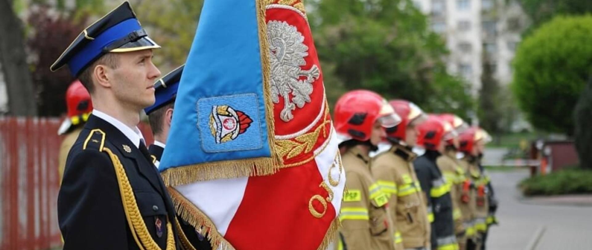 strażacy stojący w szeregu, dwóch pierwszych w umundurowaniu wyjściowym, drugi trzyma sztandar, pozostali w ubraniach bojowych i czerwonych hełmach 