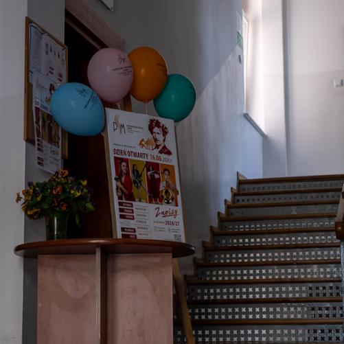 Korytarz szkoły z perspektywy wejścia głównego do budynku szkoły muzycznej. Stolik z kwiatami, za stolikiem sztaluga z plakatem informującym o dniu otwartym. Na górze plakatu doklejone baloniki z logo szkoły muzycznej.