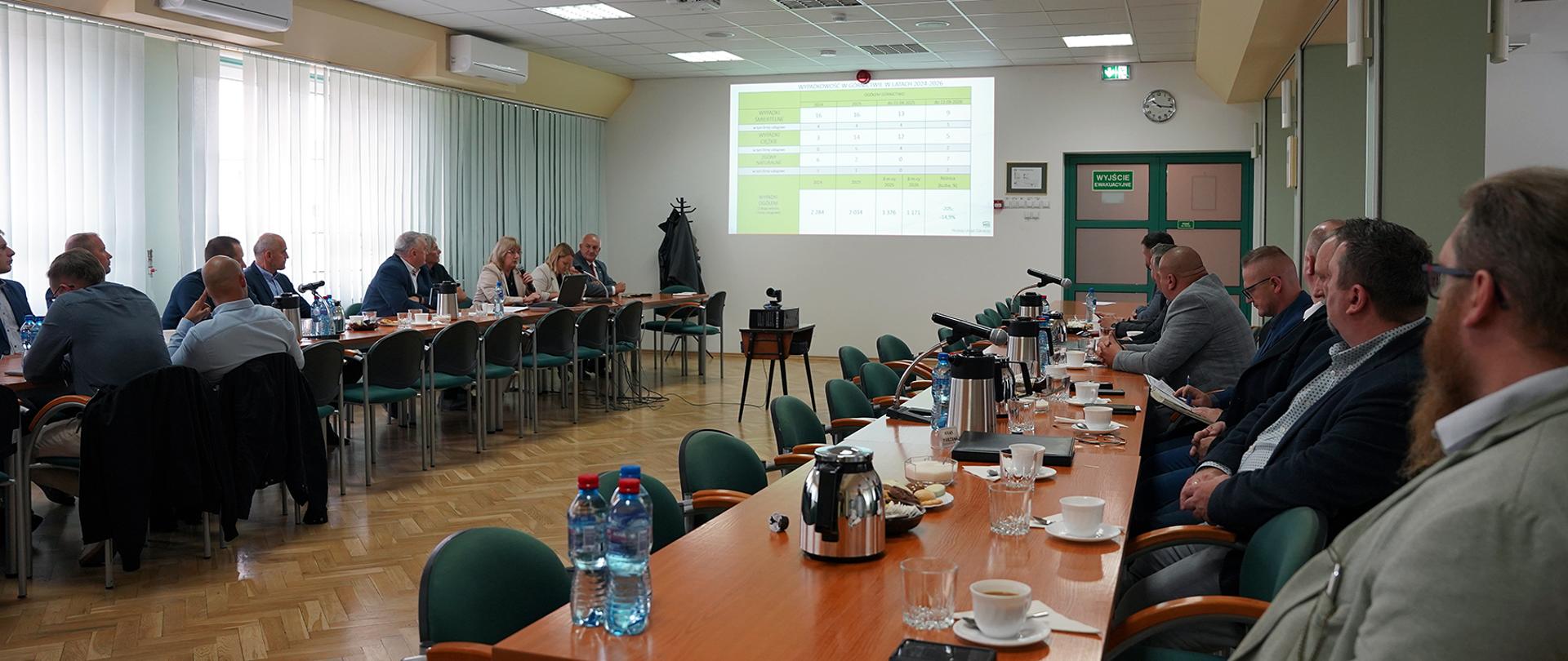 Grupa osób siedzi przy dwóch podłużnych stołach. Ich wzrok jest skierowany na ekran, na którym wyświetlana jest prezentacja.