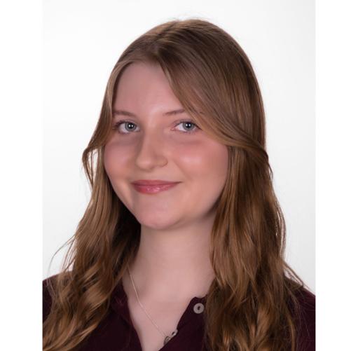 Zdjęcie kolorowe. Portret uśmiechniętej kobiety o jasnej karnacji, zielonych oczach i długich, pofalowanych włosach w kolorze jasnego brązu z blond refleksami. Kobieta ma delikatny makijaż, lekko błyszczące usta i delikatny łańcuszek na szyi. Ubrana w ciemnobordową koszulkę polo z rozpiętymi guzikami. Tło jednolite, białe.
