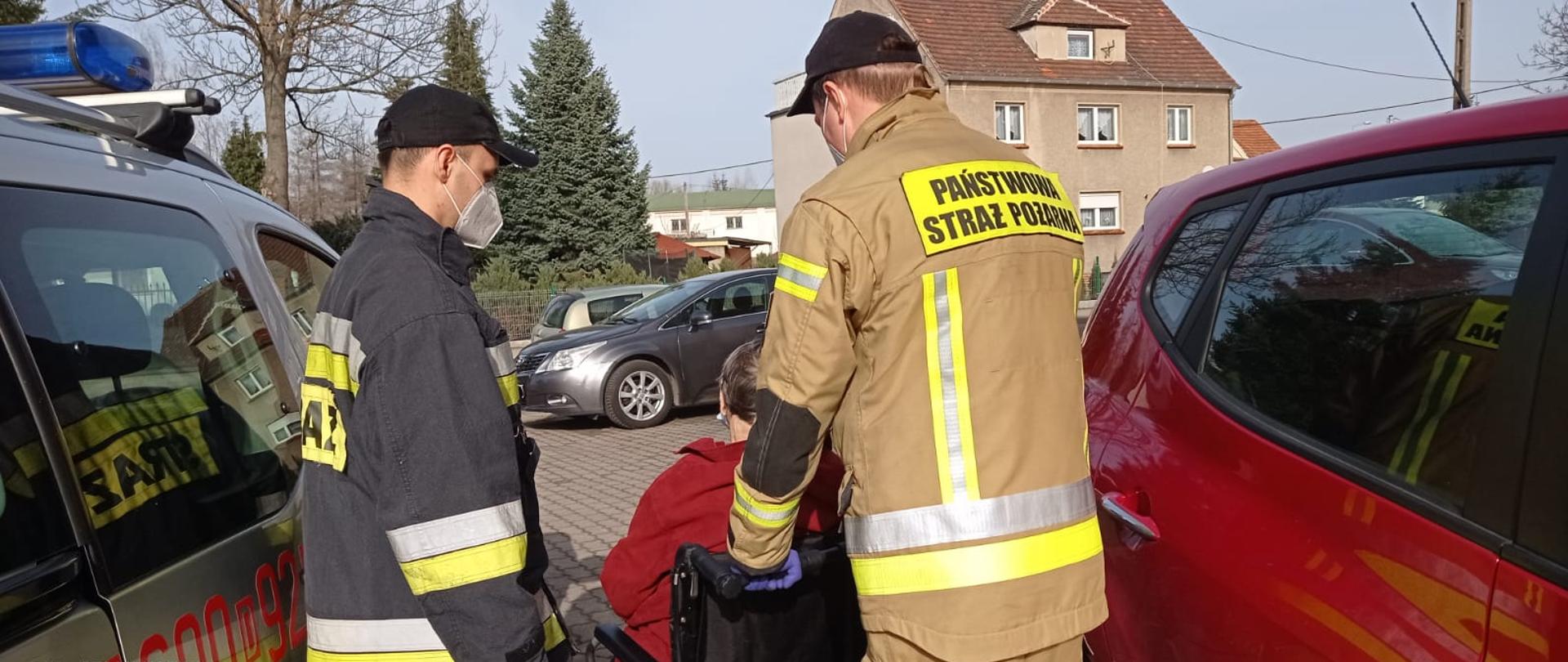 Dwóch funkcjonariuszy PSP pomaga starszej osobie poruszającej się na wózku inwalidzkim w dotarciu do punktu szczepień.