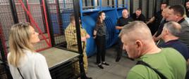 Na zdjęciu widać strażaka PSP w umundurowaniu piaskowym oraz zaproszonych gości wraz z Panią tłumacz, rozmawiają na temat przeprowadzanych ćwiczeń w komorze dymowej. Z tyłu widać elementy toru przeszkód w komorze dymowej.