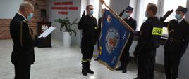 na zdjęciu w sali konferencyjnej komendy, ślubowanie dwóch nowo przyjętych strażaków na sztandar KP PSP w Łowiczu. słowa roty odczytuje komendant powiatowy PSP w Łowiczu mł. bryg. Arkadiusz Makowski, ubrany w mundur galowy ze sznurem bez nakrycia głowy, funkcjonariusze trzymają prawą rękę w geście przysięgi, strażacy ubrani w ubranie koszarowe i czarne rękawiczki bez nakrycia głowy. Trzyosobowy poczet sztandarowy salutujący na sztandar, ubrany w mundur galowy ze sznurem i nakryciem głowy w postaci rogatywki. Zdjęcie w formacie panoramy.