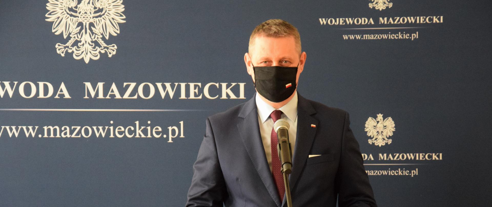 Elegancko ubrany mężczyzna przemawia podczas uroczystości wręczania odznaczeń państwowych. W tle widać granatową ściankę z białymi napisami