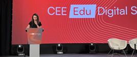 Ubrana na ciemno kobieta stoi przy mównicy i mówi do mikrofonu, za nią czerwona ściana z napisem CEE Edu Digital Summit.