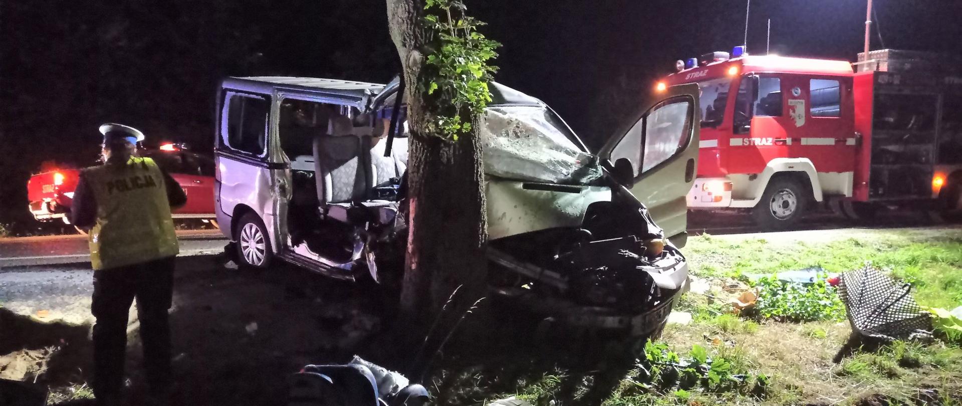Zdjęcie przedstawia uszkodzony w wyniku zderzenia samochód osobowy typu bus stojący w przydrożnym rowie, który uderzył przodem w drzewo oraz samochód pożarniczy i policjantkę.
W tle jezdnia, drugi samochód pożarniczy oraz las.
