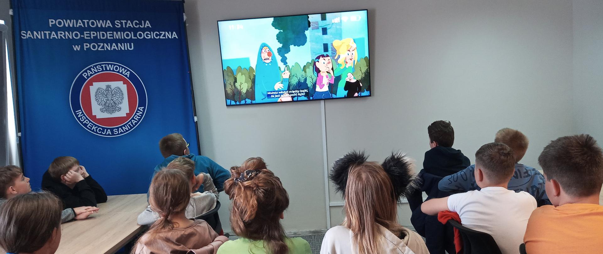 Dziewczęta i chłopcy w wieku 11 lat, siedzący na krzesłach, przy stołach. Zwróceni twarzami w kierunku ekranu umieszczonego na ścianie, po środku sali. Na ekranie kadr z filmu edukacyjnego, ukazujący postaci dwóch dziewcząt i chłopaka, który pali przy nich papierosa. Sala o jasnych ścianach. Po lewej stronie od ekranu monitora duży baner z granatowym tłem, logo Państwowej Inspekcji Sanitarnej (biały orzeł na tle czerwonego okręgu) i napisem „Powiatowa Stacja Sanitarno-Epidemiologiczna w Poznaniu”. 