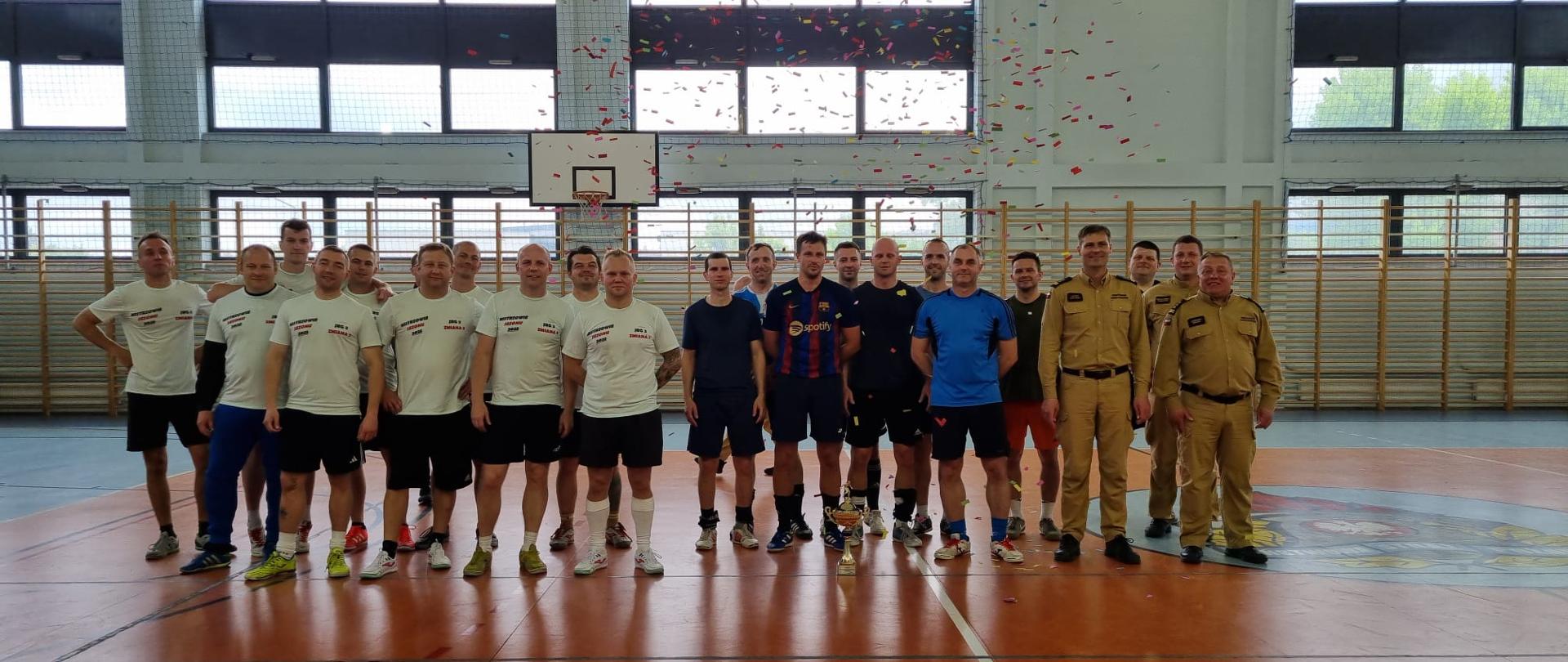 Grupa mężczyzn na hali sportowej podczas uroczystego podsumowania turnieju. Część w strojach sportowych, część w mundurach strażackich. Na ziemi stoi puchar, a w powietrzu unosi się kolorowe konfetti.
