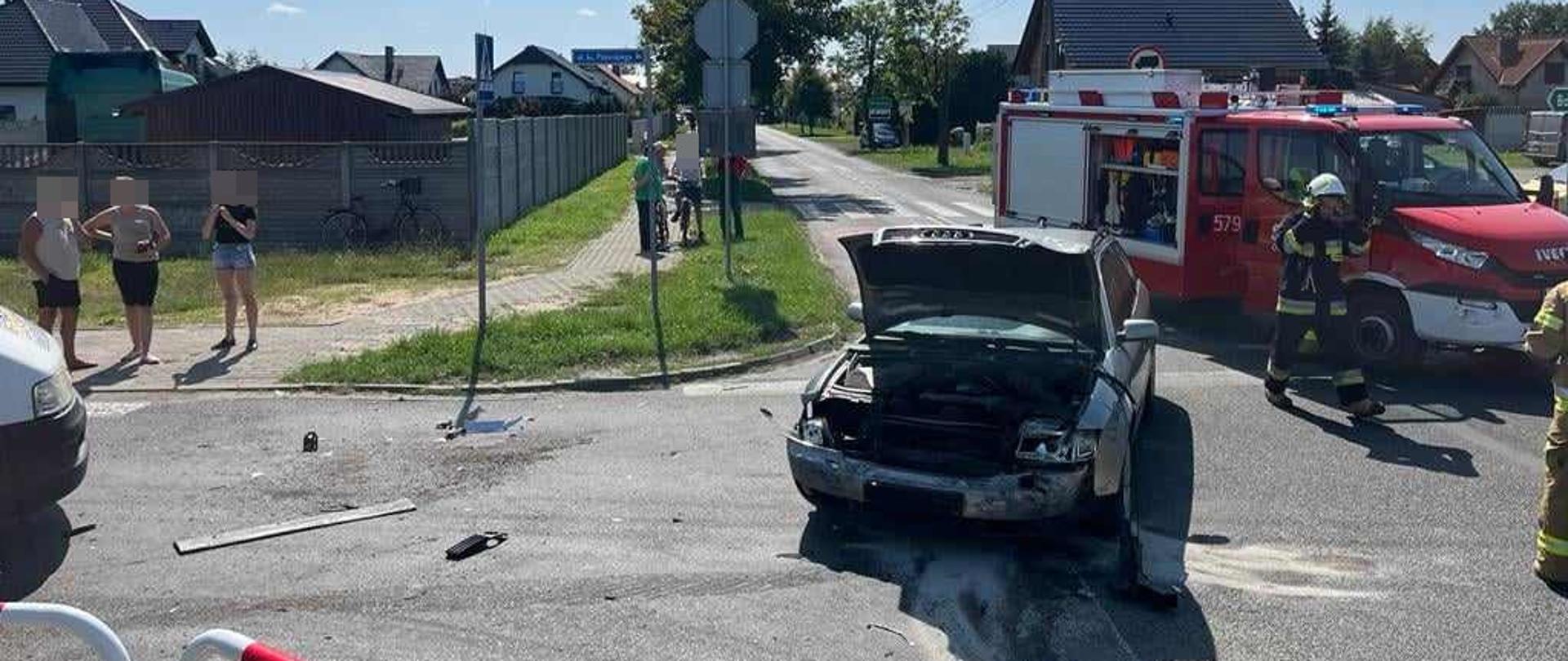Zdjęcie przedstawia skrzyżowanie dróg. Na nim jeden z pojazdów biorących udział w zderzeniu. Jest to samochód osobowy koloru srebrnego. W aucie osobowym podniesiona jest pokrywa silnika. Za nim stoi wóz strażacki. Obok widoczny jest strażak w białym hełmie. Z lewej strony na chodniku stoją 3 osoby.