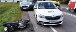 Na jezdni stoi samochód w kolorze białym po zderzeniu z motocyklem, na poboczu drogi leży uszkodzony wrak motocykla. W oddali zdjęcia widoczne pojazdy Policji i Karetki Pogotowia. 