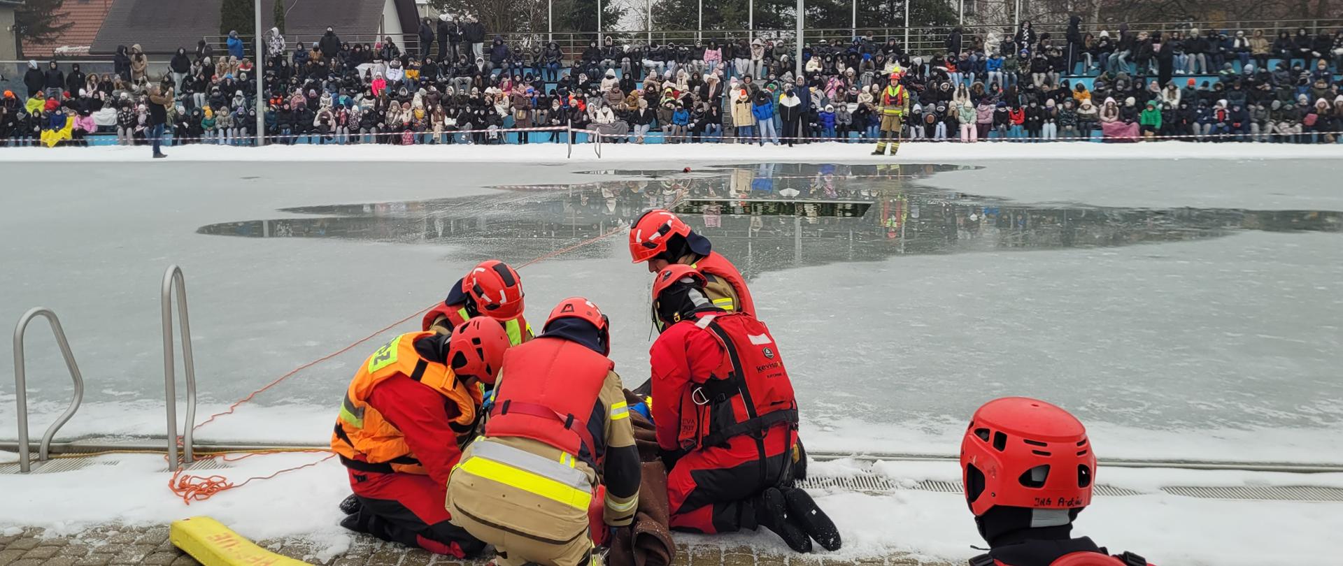 Grupa strażaków na brzegu zamarzniętego basenu udziela pomocy poszkodowanemu. Część strażaków ubrana w skafandry do pracy w wodzie. W tle widoczna widownia.