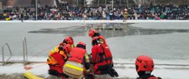 Grupa strażaków na brzegu zamarzniętego basenu udziela pomocy poszkodowanemu. Część strażaków ubrana w skafandry do pracy w wodzie. W tle widoczna widownia.