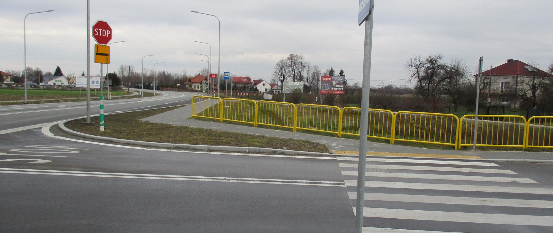 Przejście dla pieszych przy skrzyżowaniu - czerwony znak "stop" przed skrzyżowaniem i żółta tablica ze schematem skrzyżowania, znak "uwaga przejście dla pieszych", żółte barierki oddzielające chodnik od jezdni za przejściem dla pieszych, w oddali domy 