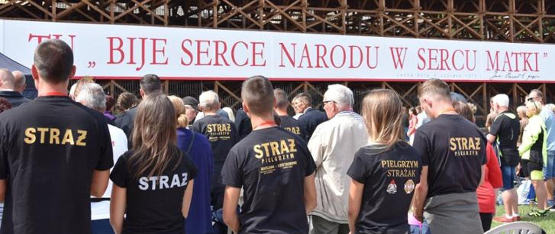 Strażacy na pielgrzymce stojący tyłem w koszulkach z napisem straż. 