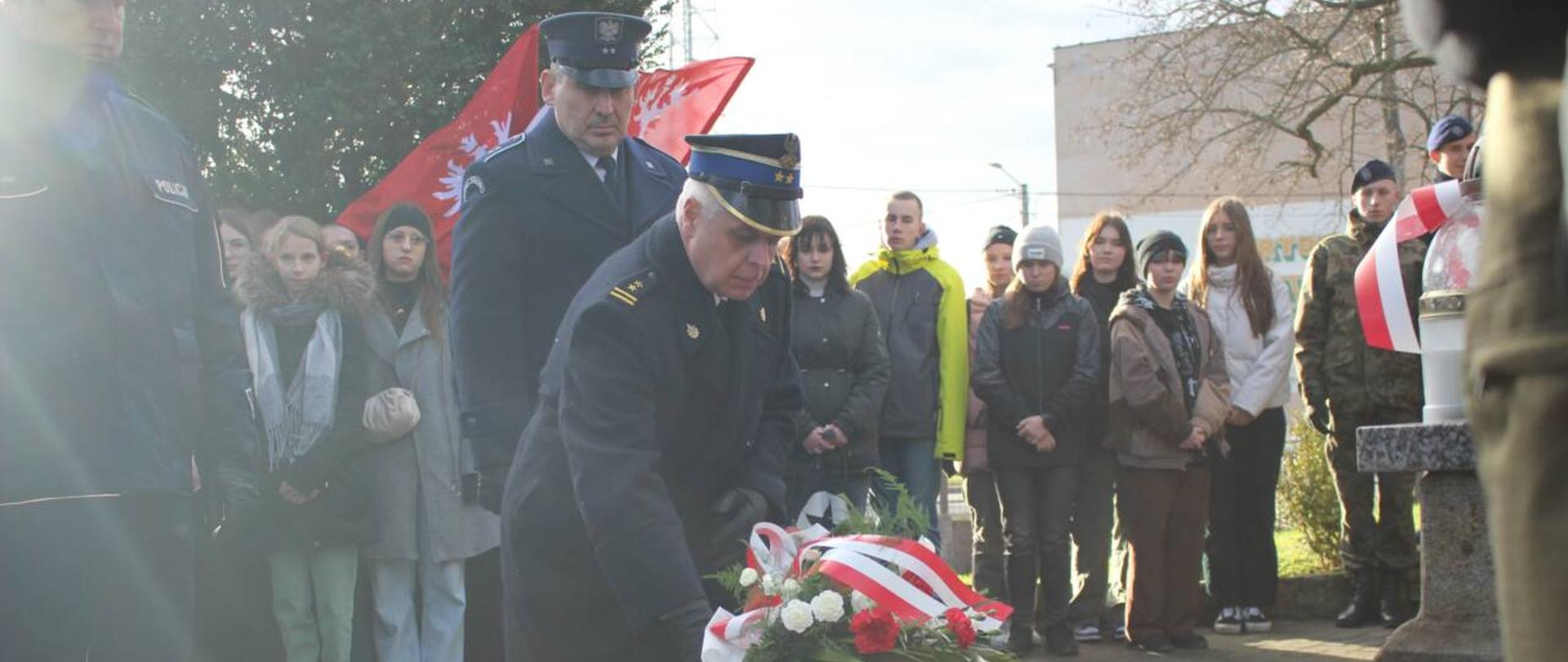 104 rocznica Zwycięskiego Powstania Wielkopolskiego. Cmentarz parafialny w Rawiczu. Zastępca komendanta powiatowego PSP w Rawiczu składa wiązankę kwiatów pod obeliskiem ku czci Powstańców Wielkopolskich. Towarzyszą mu funkcjonariusz policji i służby więziennej. Kwiaty w wiązance mają kolor biały i czerwony. Ponadto wiązanka udekorowana jest biało-czerwoną wstążką. W tle stoją pozostali uczestnicy uroczystości.