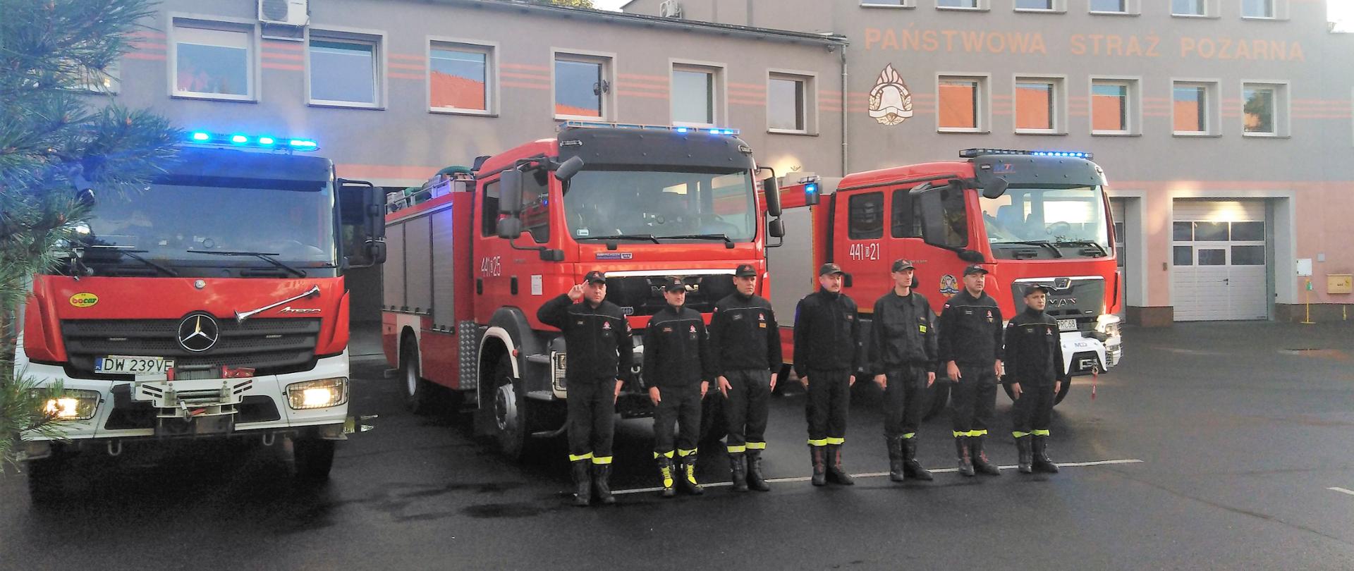 Obraz przedstawia strażaków JRG Jawor podczas minuty ciszy dla zmarłego strażaka z OSP, który zginął podczas prowadzenia działań ratowniczych. Straży w umundurowaniu koszarowym na tle pojazdów pożarniczych i budynku komendy.