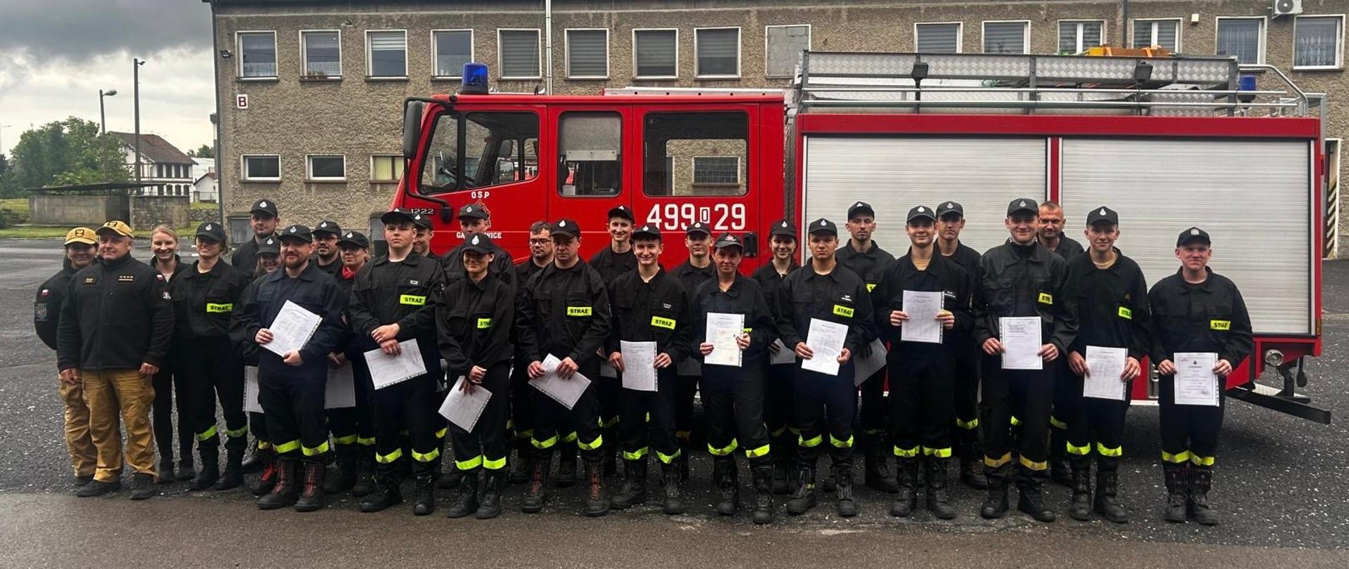 Na zdjęciu znajdują się strażacy w umundurowaniu koszarowym na tle samochodu bojowego trzymający w rękach zaświadczenia z ukończenia szkolenia