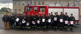 Na zdjęciu znajdują się strażacy w umundurowaniu koszarowym na tle samochodu bojowego trzymający w rękach zaświadczenia z ukończenia szkolenia
