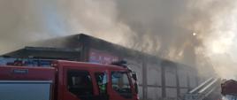 Zdjęcie przedstawia samochód pożarniczy oraz mechaniczną drabinę pożarniczą na tle budynku magazynowego z którego wydobywają się kłęby dymu.