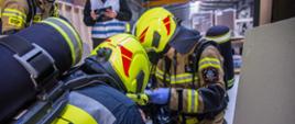 
Strażacy w ubraniach specjalnych udzielają pierwszej pomocy osobie poszkodowanej podczas ćwiczeń w budynku produkcyjnym. W tle strażak w kamizelce ostrzegawczej oceniający działania. W tle widać użyte opatrunki.