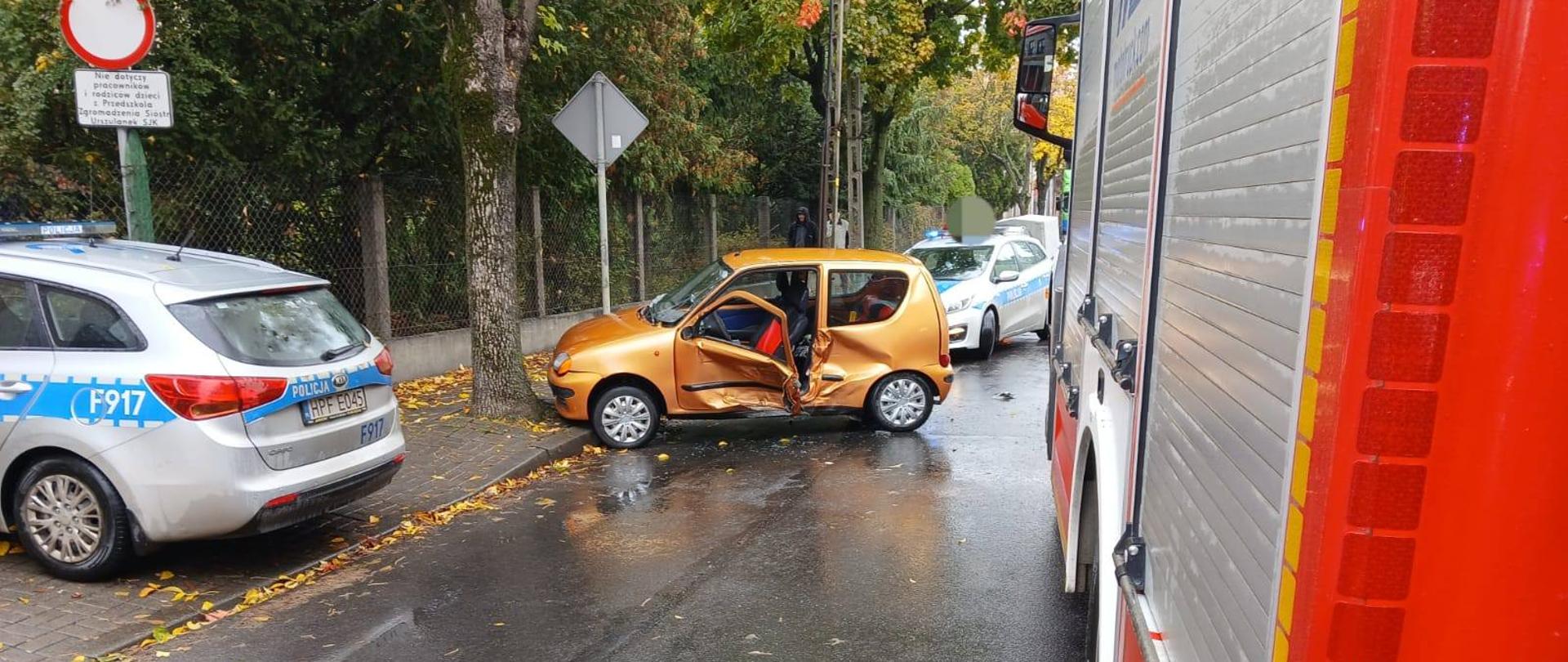 Na zdjęciu widać miejsce wypadku drogowego z udziałem pomarańczowego samochodu osobowego. Pojazd stoi częściowo na jezdni i chodniku, z wyraźnie wgniecionymi drzwiami po lewej stronie. Drzwi kierowcy są otwarte. Obok pojazdu znajduje się radiowóz policyjny, a po prawej stronie zdjęcia widoczny jest czerwony wóz straży pożarnej. Jezdnia jest mokra. W tle widać drzewa i znaki drogowe.